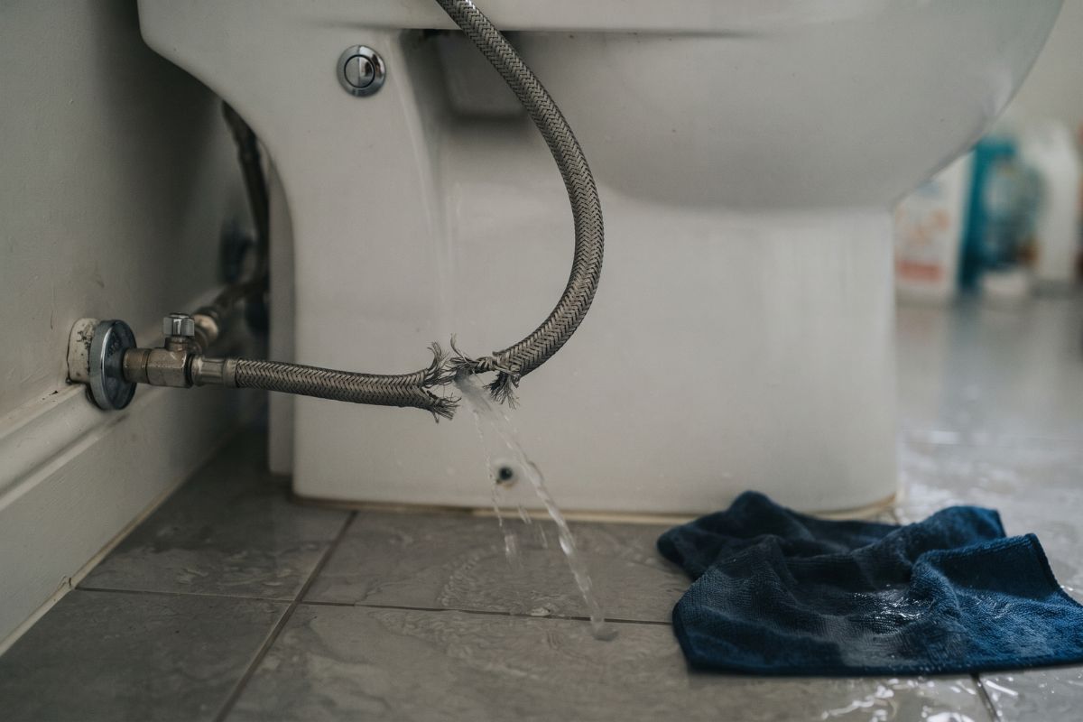 Split braided flexible hose leaking water onto a bathroom floor beside a toilet

